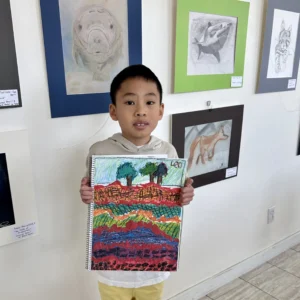 Child holding a colourful landscape artwork at Aliki’s Art House student art show.