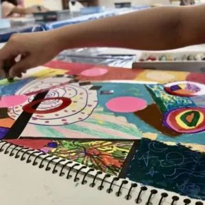 Child creating a colorful abstract mixed-media artwork in a sketchbook at Aliki’s Art House.